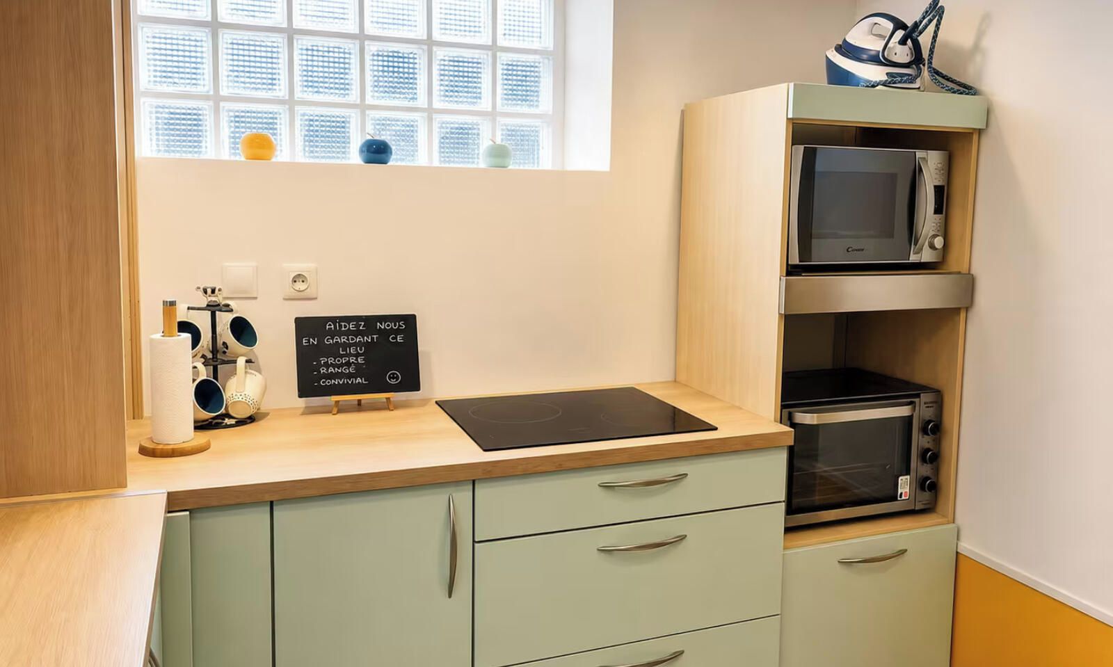 Une petite cuisine équipée avec une plaque de cuisson à induction noire, un four à micro-ondes et un four grille-pain. On y trouve également un tableau noir avec du texte, une fenêtre avec des stores et une petite étagère avec des objets décoratifs.
