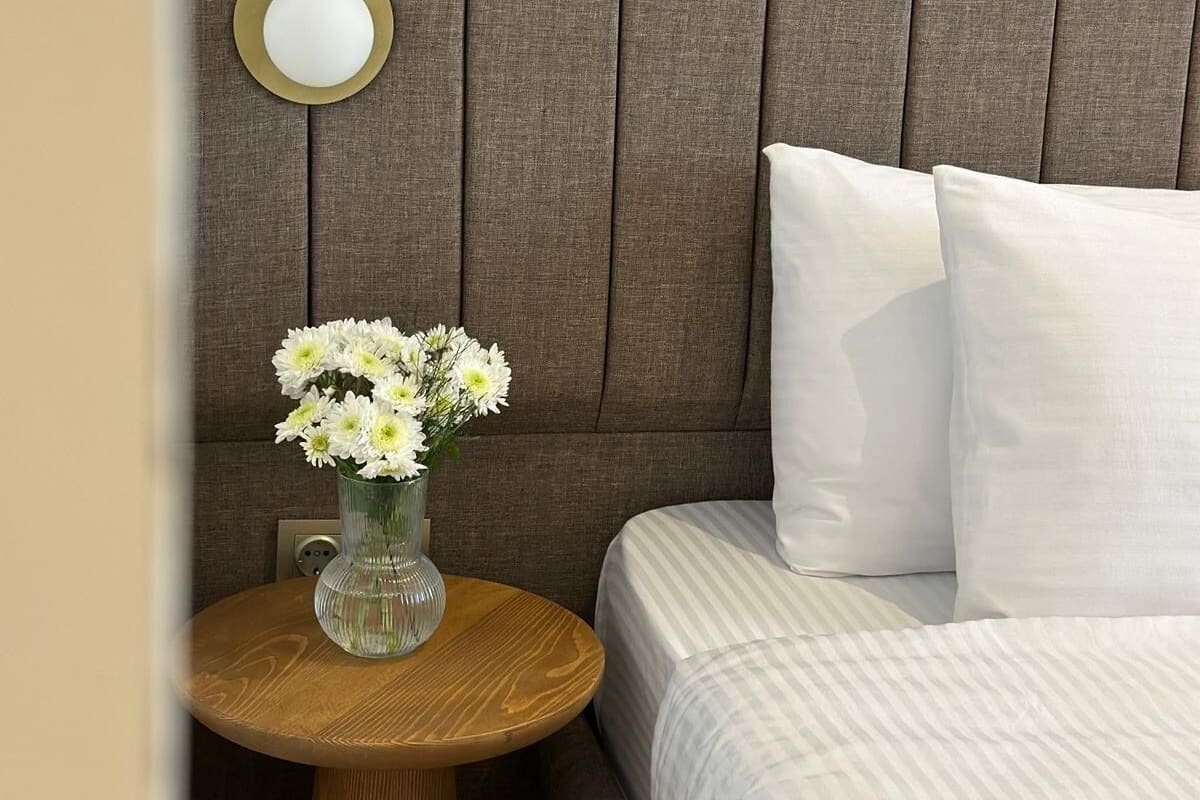 A bedroom with a bed, pillows, a round wooden nightstand, and a vase with flowers.