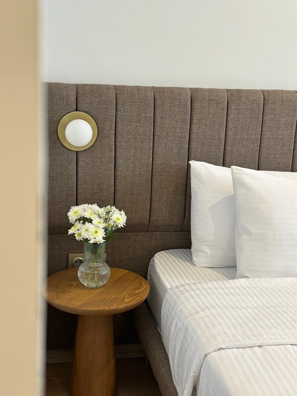 Une chambre à coucher avec un lit, des oreillers, une table de chevet ronde en bois et un vase rempli de fleurs.