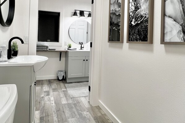 A modern bathroom with a white sink, black faucet, and a mirror above it. There are framed pictures on the wall and a window with curtains.