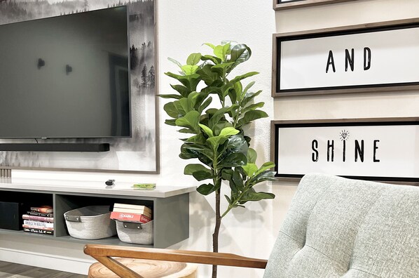 A living room with a flat-screen TV, a bookshelf with books and decorative items, a plant, and framed motivational quotes on the wall.