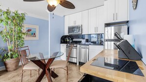 A modern kitchen with white cabinets, stainless steel appliances, a dining table with chairs, and a ceiling fan.