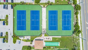 An aerial view of a sports complex with two tennis courts, parking areas, and a small building with a sign.