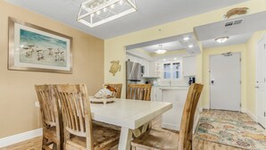 A dining area with a white table and wooden chairs, a framed artwork on the wall, and a kitchen in the background.