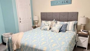 Un dormitorio con cama, almohadas, una mesita de noche, una lámpara, un letrero en la pared y una puerta.