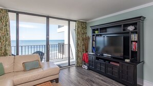 A living room with a large TV, a sofa, and a view of the ocean through a sliding glass door.