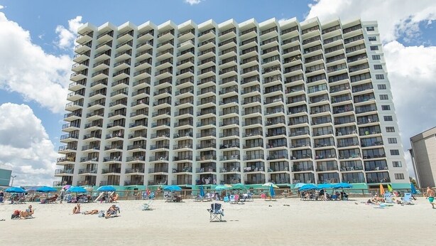 A high-rise hotel with multiple balconies, a sandy beach in front, and beach umbrellas.