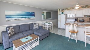 Un salon avec un canapé gris, une table basse et un tableau représentant l’océan. On y trouve également un coin repas avec une table et des chaises, ainsi qu’une cuisine dotée de meubles blancs et d’appareils électroménagers.