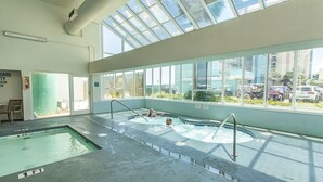 Une grande piscine intérieure aux parois en verre transparent, avec un espace peu profond séparé et une fenêtre panoramique donnant sur un parking.