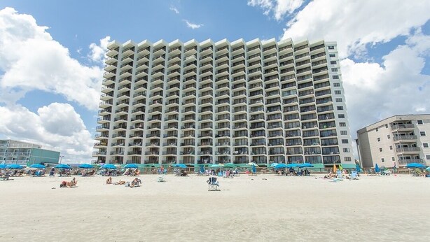 Un hôtel de grande hauteur doté de nombreux balcons, situé en bord de plage, avec du sable fin et des parasols bleus.