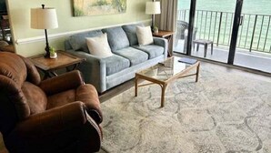 A living room with a sofa, armchair, and coffee table, a painting on the wall, and a view of the ocean through the balcony doors.