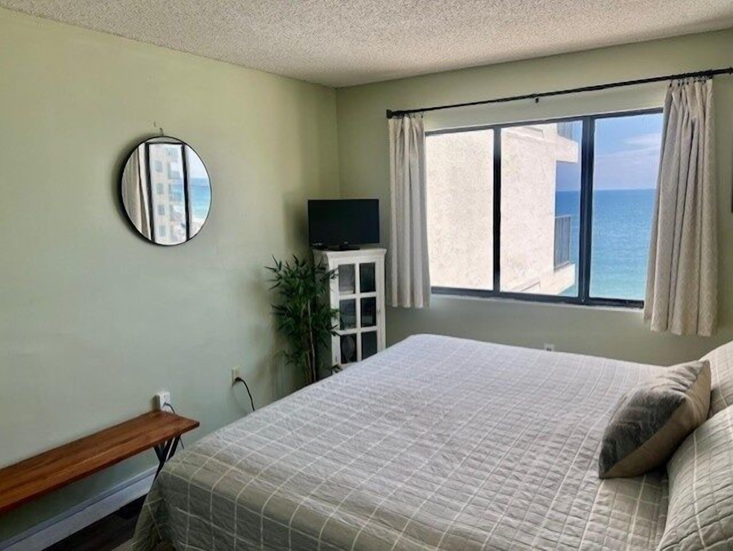 A bedroom with a bed, a television on a cabinet, a mirror on the wall, and a window with curtains.