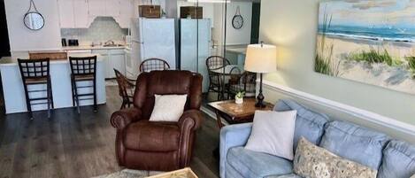 A living room with a leather armchair, a blue sofa, and a coffee table. There is a painting on the wall and a kitchen area in the background.