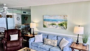 A living room with a blue sofa, a brown leather chair, and a glass coffee table. There is a painting of a beach on the wall.