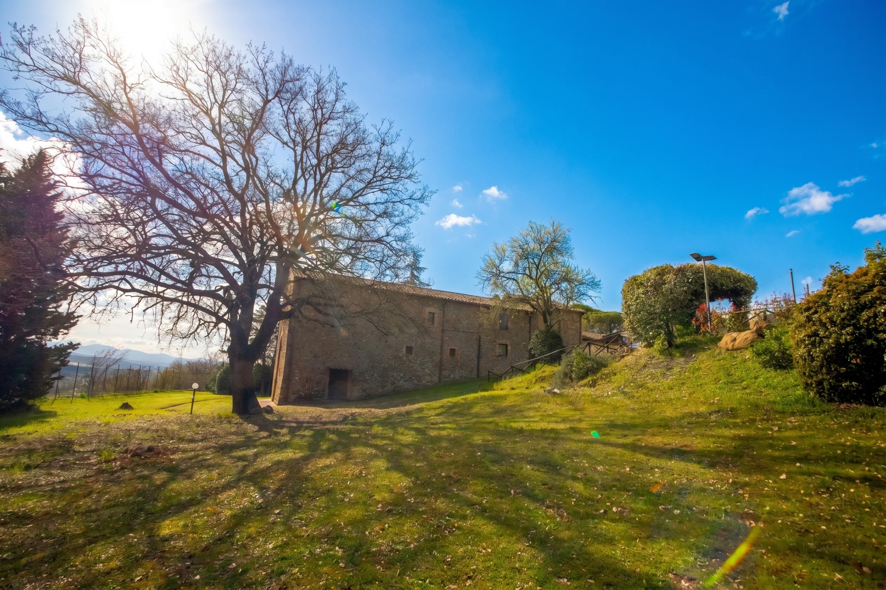 Edifici de pedra amb teulada inclinada, envoltat d'una zona verda amb arbres i un cel serè.
