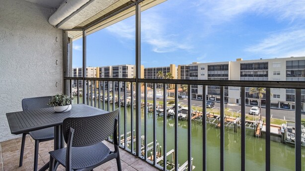Un balcon avec une table et des chaises, donnant sur un canal où l’on aperçoit des bateaux et des bâtiments en arrière-plan.