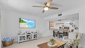 Un vaste salon doté d’un ventilateur de plafond, d’un grand téléviseur, d’un coin repas avec des chaises et d’une cuisine à l’arrière-plan.