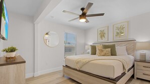 A bedroom with a bed, bedside tables, a ceiling fan, a TV, a mirror, and framed pictures on the wall. - Welcome To Coconut Dreams Fantastic Vacation Rental - 2633 Estero Blvd Unit A 3 Bedroom Home by RedAwning (Fort Myers Beach)