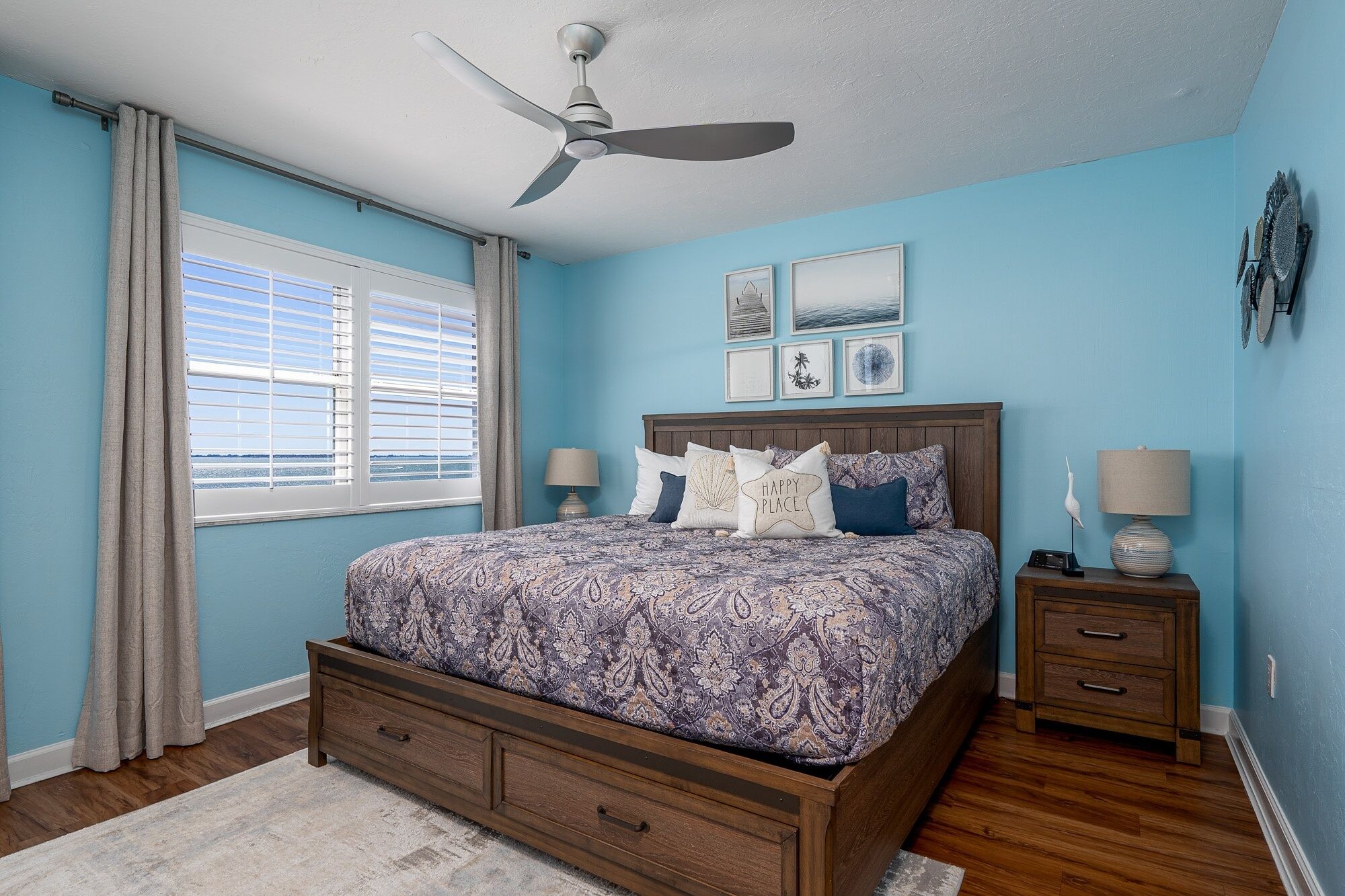 Quarto com cama, duas mesas de cabeceira, ventilador de teto e janela com cortinas.