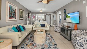 A spacious living room with a high ceiling fan, a large flat-screen TV, and a dining area in the background.