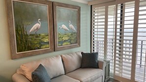 Un salón con un sofá color beige, dos cuadros enmarcados de aves y una ventana con persianas.