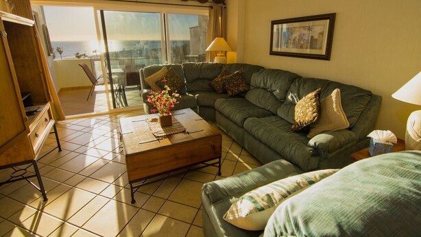 A living room with a green sofa, a wooden coffee table with a vase of flowers, a lamp, and a view of the sea through large windows. - Luxurious 1 Bedroom Condo on Sandy Beach at Las Palmas Resort B-402 1 Condo by RedAwning (Puerto Peñasco)
