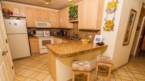 A kitchen with a granite countertop, two bar stools, a refrigerator, a microwave, an oven, and a sink. - Luxurious 1 Bedroom Condo on Sandy Beach at Las Palmas Resort B-402 1 Condo by RedAwning (Puerto Peñasco)