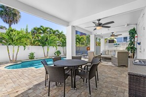 Un patio exterior con piscina, muebles de mimbre, ventiladores de techo y vista a un edificio cercano.