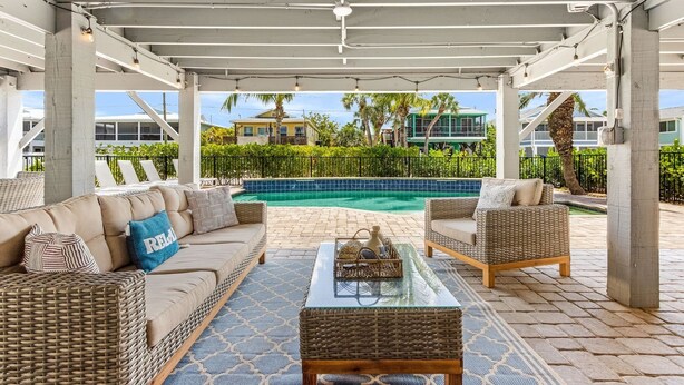 A covered outdoor seating area with wicker furniture, a glass coffee table, and a view of a swimming pool. - Shells N Sunshine! Getaway Destination Just Minutes From The White Sandy Beaches Of Fort Myers! 4 Bedroom Home by RedAwning (Fort Myers Beach)