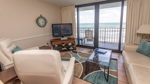 Una sala de estar con televisor, dos sofás, una mesa de centro y vista al océano a través de amplios ventanales correderos.