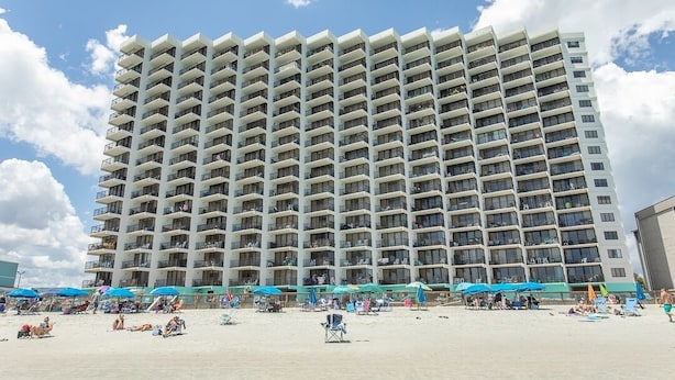 Un hotel de gran altura con varios balcones, una playa de arena frente a él y sombrillas de playa.