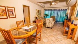 A dining area with a glass-top table and wooden chairs, a sofa, and a television in a living room with a ceiling fan. - Spectacular 2 Bedroom Condo on Sandy Beach at Las Palmas Resort B-203 2 Condo by RedAwning (Puerto Peñasco)