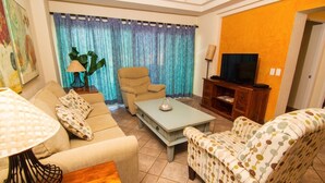 A living room with a sofa, armchair, coffee table, TV stand, and curtains. - Spectacular 2 Bedroom Condo on Sandy Beach at Las Palmas Resort B-203 2 Condo by RedAwning (Puerto Peñasco)