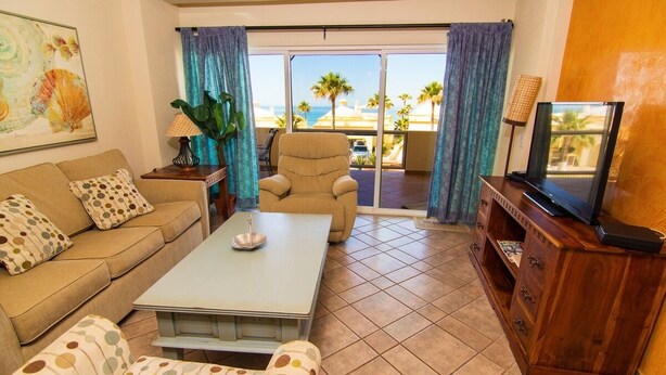 A living room with a sofa, armchair, and coffee table, a wooden cabinet with a TV, and a balcony with a view of palm trees and the ocean. - Spectacular 2 Bedroom Condo on Sandy Beach at Las Palmas Resort B-203 2 Condo by RedAwning (Puerto Peñasco)