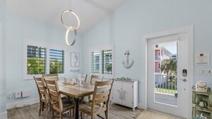 A dining area with a wooden table and chairs, a white cabinet with decorative items, and a view of a building through the window. - Welcome To Mermaid Bay Vacation Rental - 261 Delmar 3 Bedroom Home by RedAwning (Fort Myers Beach)