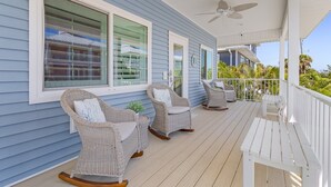 A covered porch with wicker rocking chairs and a bench, large windows, and a ceiling fan. - Welcome To Mermaid Bay Vacation Rental - 261 Delmar 3 Bedroom Home by RedAwning (Fort Myers Beach)