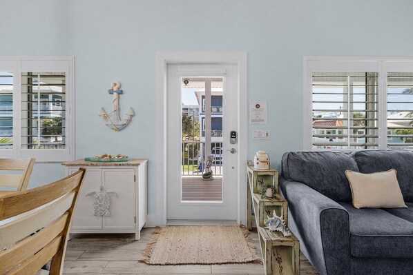 Een gezellige woonkamer met een grijze bank, een houten stoel en een kleine tafel met decoratieve objecten. Een witte deur leidt naar een balkon met uitzicht op gebouwen en een blauwe auto.