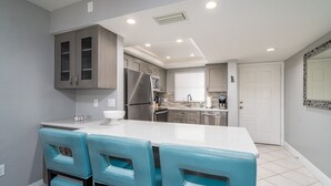A modern kitchen with grey cabinets, a stainless steel refrigerator, and a white countertop. There are three turquoise bar stools and a white bowl on the counter. - Estero Cove 132 - Beautifully Updated!! 2 Bedroom Condo by RedAwning (Fort Myers Beach)