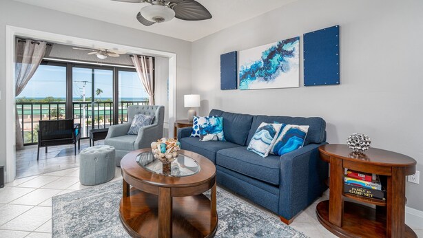 A living room with a blue sofa, a glass coffee table, and a ceiling fan. There is a view of the outdoors through large windows. - Estero Cove 132 - Beautifully Updated!! 2 Bedroom Condo by RedAwning (Fort Myers Beach)