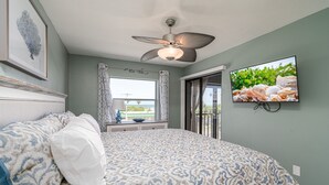 A bedroom with a bed, a ceiling fan, a TV, and a window with curtains. - Estero Cove 132 - Beautifully Updated!! 2 Bedroom Condo by RedAwning (Fort Myers Beach)