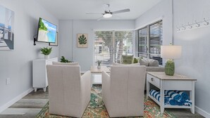 A modern living room with a ceiling fan, a flat-screen TV mounted on the wall, a small table with a plant, and a bench with a colorful rug. - 171 Washington ~ Dune Our Thing ~ Unit D ~ Fabulous Pool Home! 2 Bedroom Home by RedAwning (Fort Myers Beach)