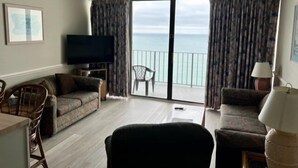 Una sala de estar con un sofá, una televisión, una mesa de comedor con sillas y vista al mar desde un balcón.