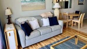 A living room with a grey sofa, a small table, and a dining area with wooden chairs. - Royal Garden Resort 904 2 Bedroom Condo by RedAwning (Murrells Inlet)