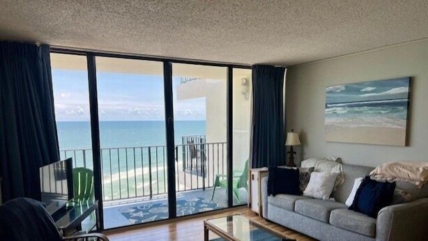 A hotel room with a balcony, a sofa, a coffee table, and a painting of a beach scene. - Royal Garden Resort 904 2 Bedroom Condo by RedAwning (Murrells Inlet)
