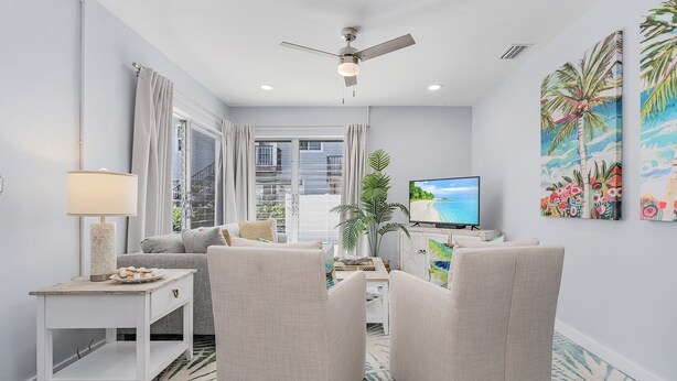 A modern living room with a sofa, armchairs, a TV, and a ceiling fan. - 171 Washington ~ Dune Our Thing ~ Unit A ~ Fabulous Pool Home! 2 Bedroom Home by RedAwning (Fort Myers Beach)