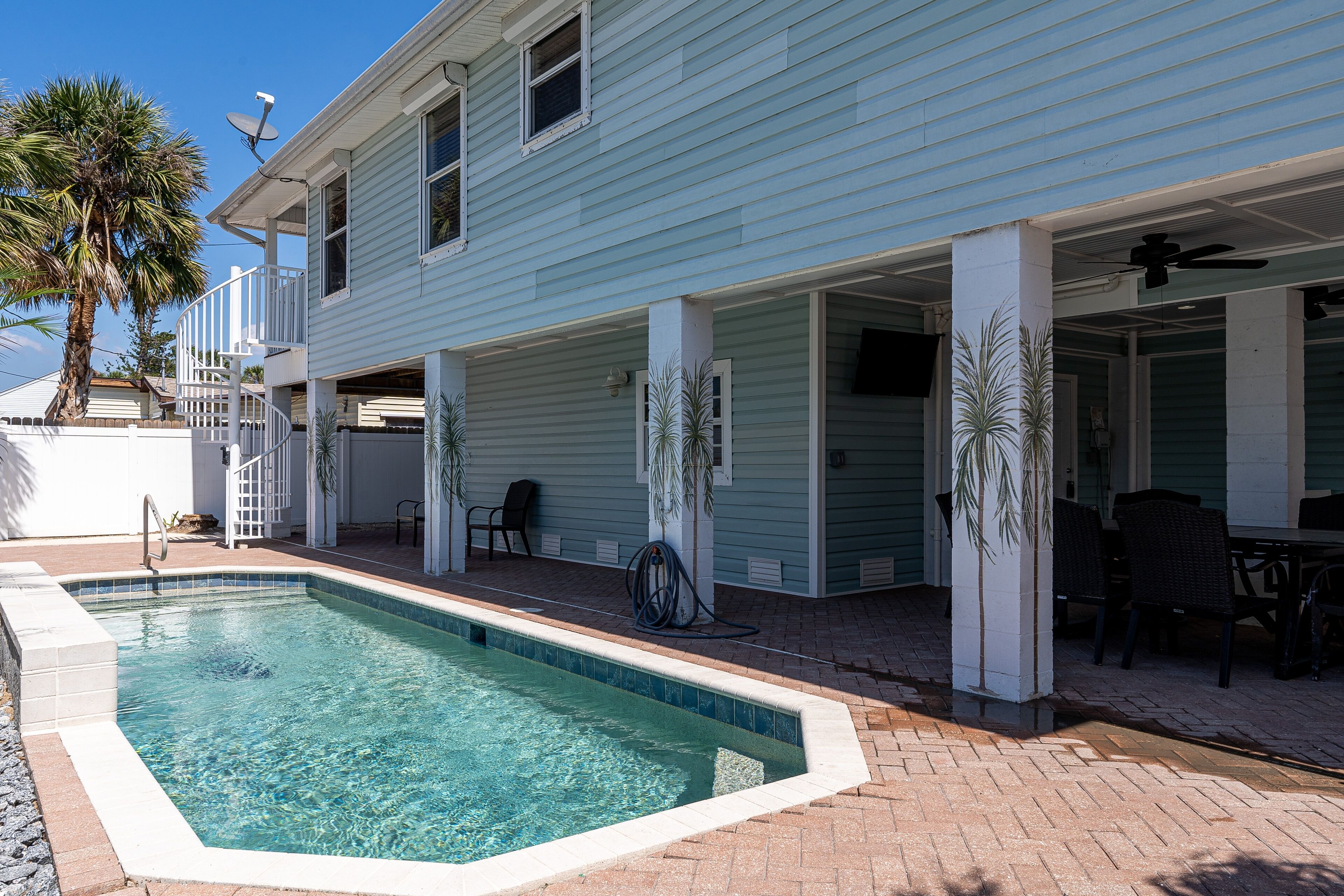 A swimming pool with a tiled deck, a covered patio area with chairs, and a palm tree.