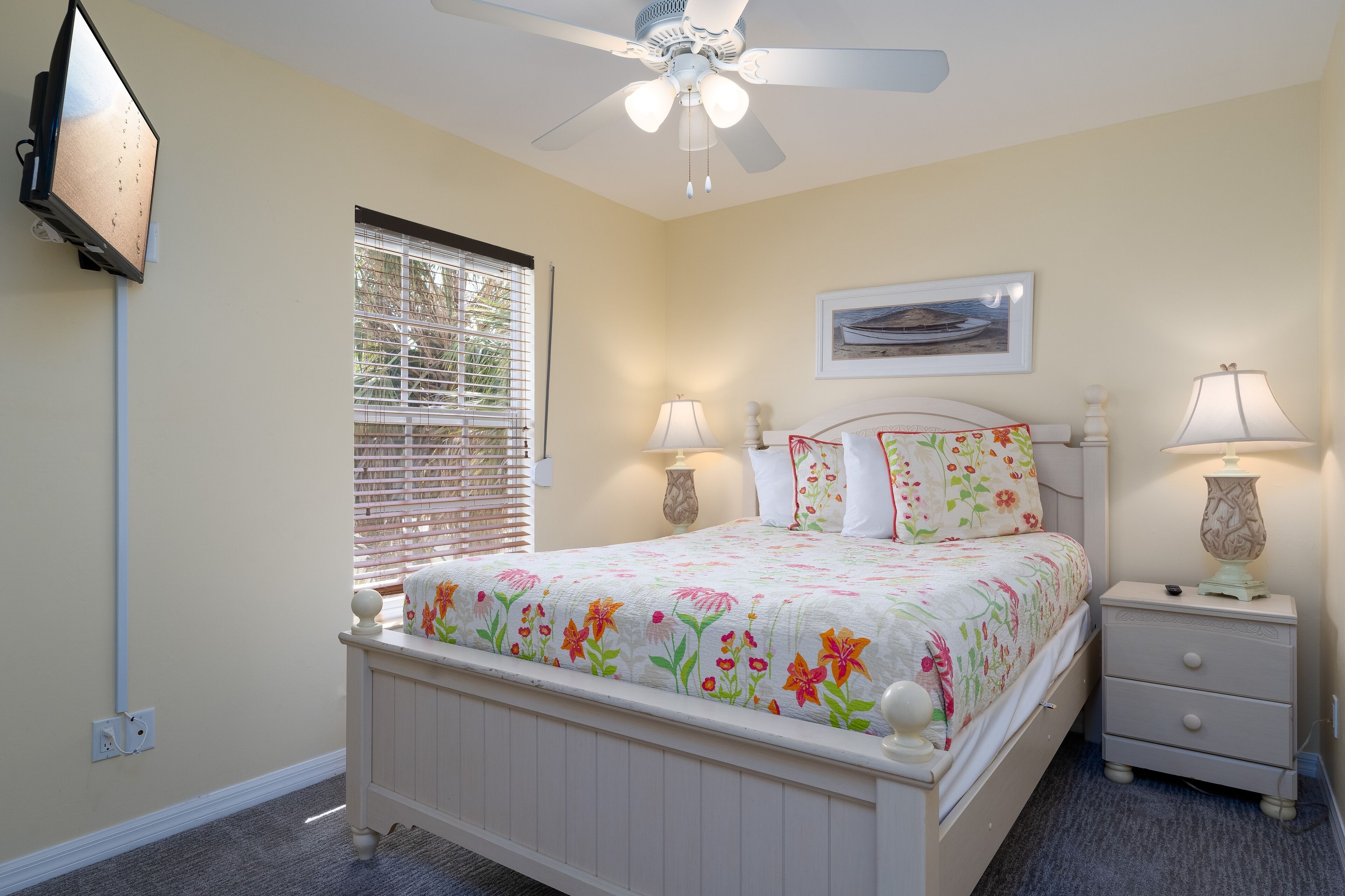 A bedroom with a bed, nightstands, a ceiling fan, a window with blinds, and a wall-mounted TV.