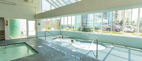 An indoor swimming pool with clear glass walls, a separate hot tub area, and a view of the cityscape through large windows.