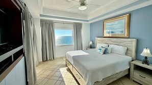 Un dormitorio con cama, ventilador de techo, ventana con cortinas, mesita de noche con lámpara y un cuadro en la pared.