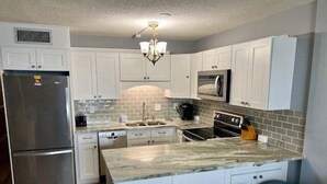 A modern kitchen with white cabinets, a stainless steel refrigerator, a built-in oven, a microwave, and a granite countertop. There are three blue bar stools at the island.